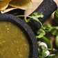 Roasted green salsa in mortar bowl with pestel, cilantro, corn chips, tomatillos, and peppers on the side