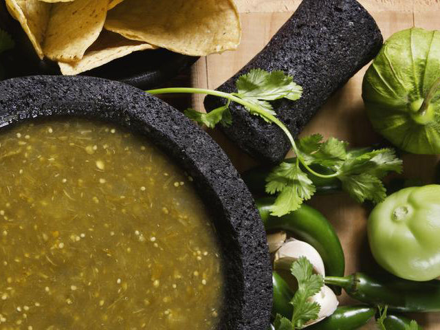 Roasted green salsa in mortar bowl with pestel, cilantro, corn chips, tomatillos, and peppers on the side